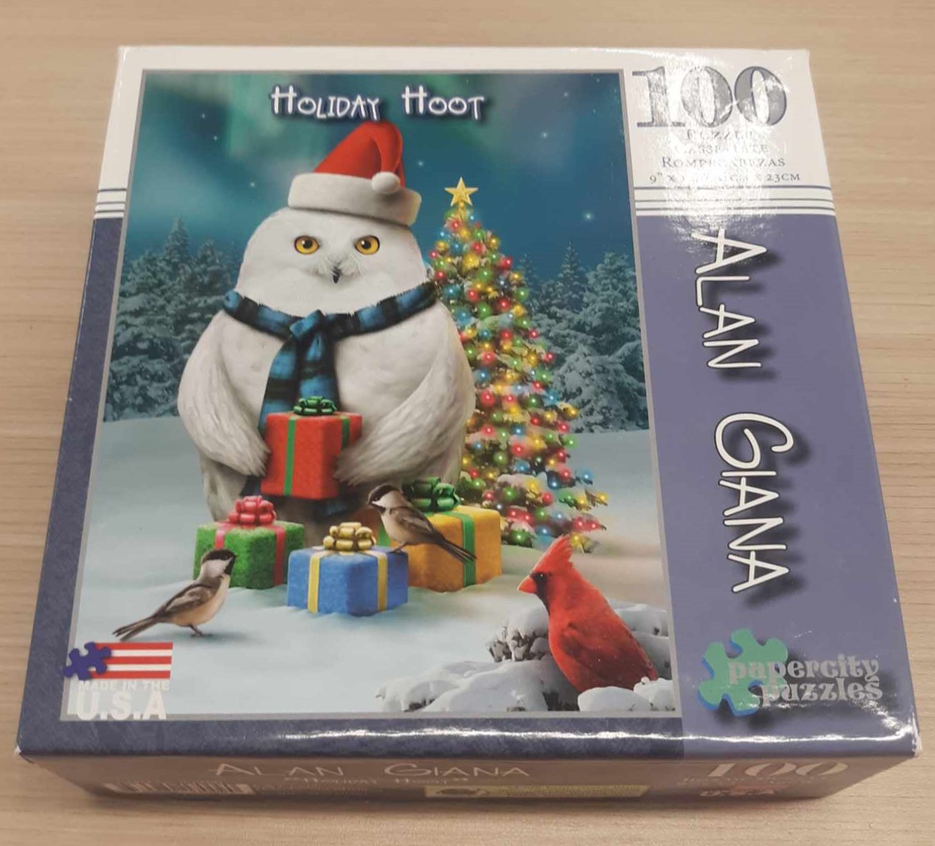 Image of a puzzle cover titled "Holiday Hoot" Showing a large owl sitting on top of presents like a nest in front of a Christmas Tree.