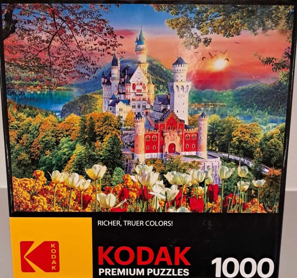 Puzzle cover showing an image of a castles at sunset near a lake with a lot of trees and flowers nearby.