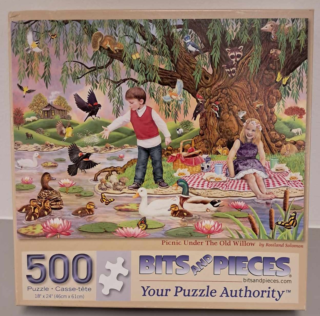 Puzzle Cover showing and image of a young boy and young girl with a picnic blanket and basket sitting under a willow tree feeding ducks in the river.