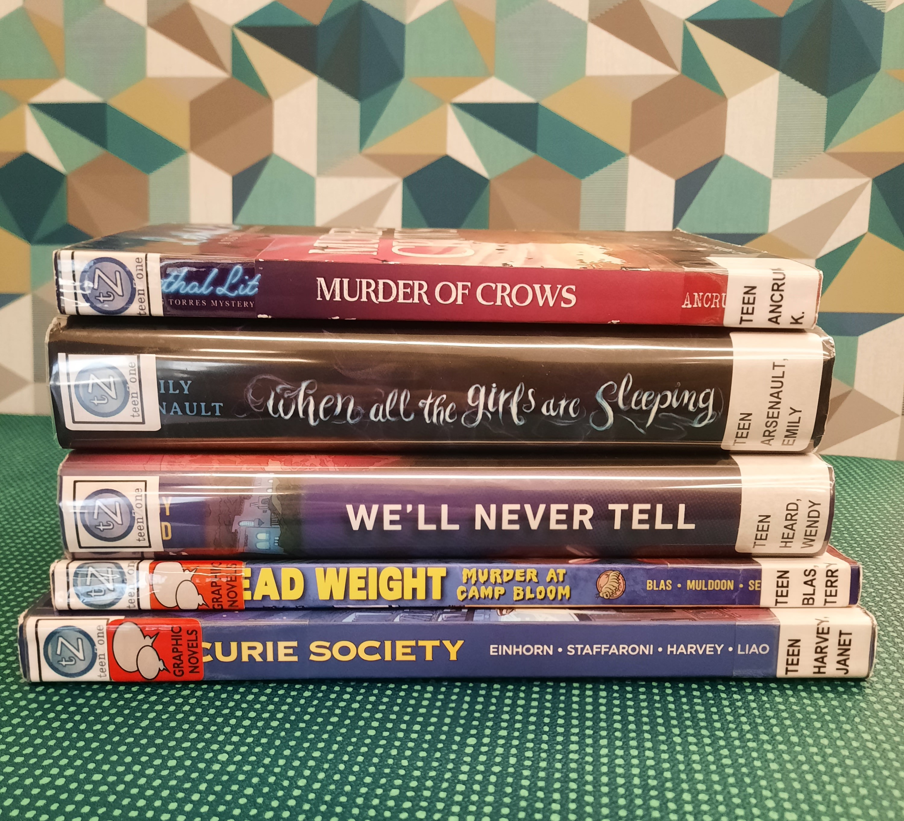 Book Stack showing the following titles: "Murder of Crows," "When All the Girls are Sleeping," "We'll Never Tell," "Dead Weight: Murder at Camp Bloom," and "The Curie Society"